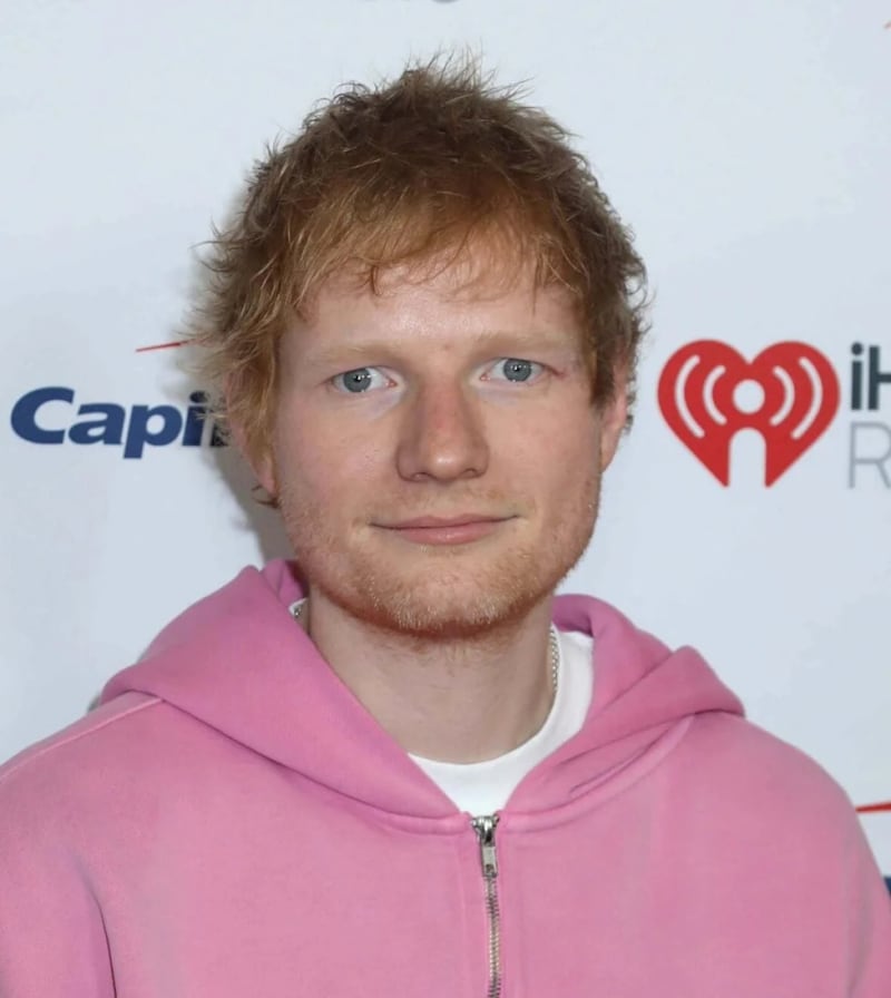 A person with reddish-brown hair wearing a pink hoodie poses in front of a Capitol and iHeartRadio branded backdrop.