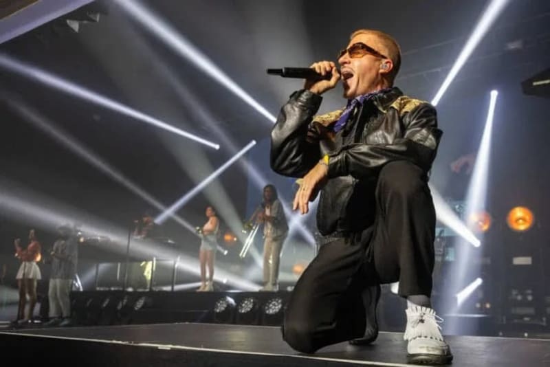 A performer in sunglasses and black jacket sings into a microphone while kneeling on stage with dramatic lighting and band members behind.