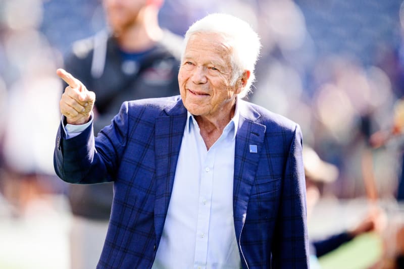 Elderly man in blue plaid blazer pointing at camera on stadium field.