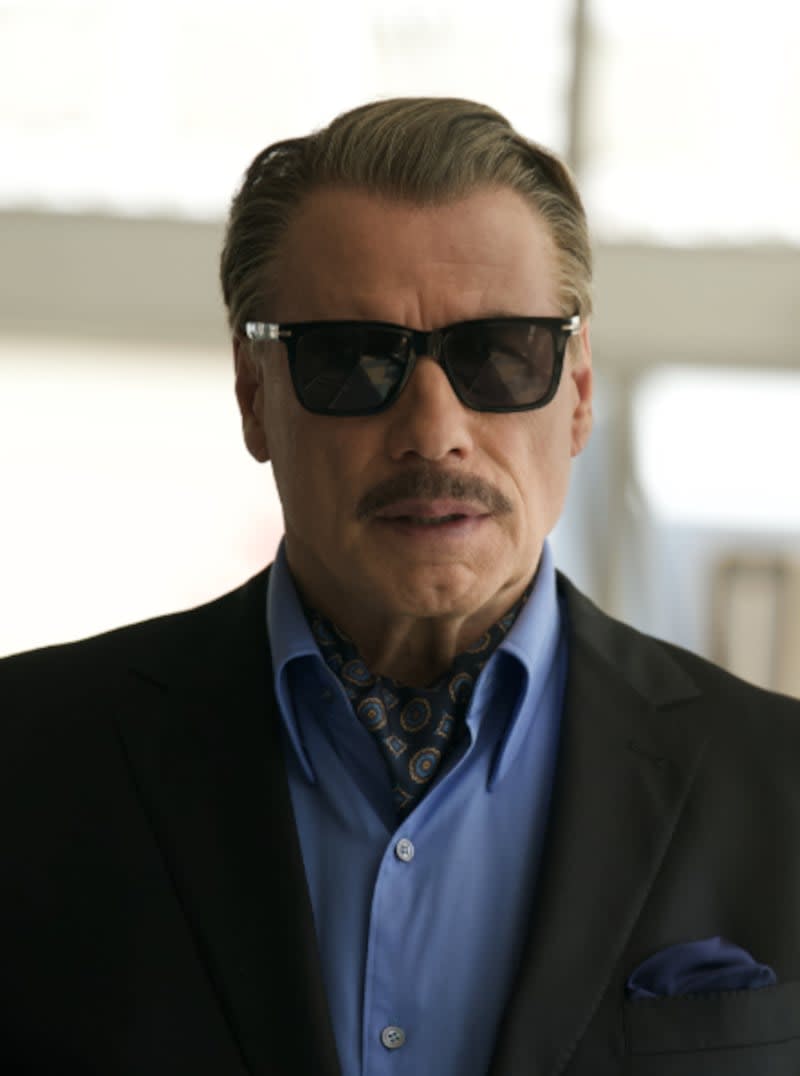 A man wearing dark sunglasses, a black blazer, and blue shirt with patterned scarf stands indoors against a light background.