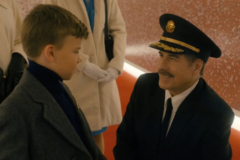 Boy in grey coat facing man in black captain's uniform and gold-badged hat, indoors.