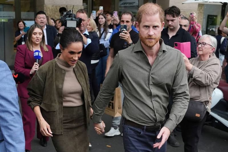 A couple in casual earth-toned clothing walks hand-in-hand through a crowded street surrounded by media personnel and onlookers.