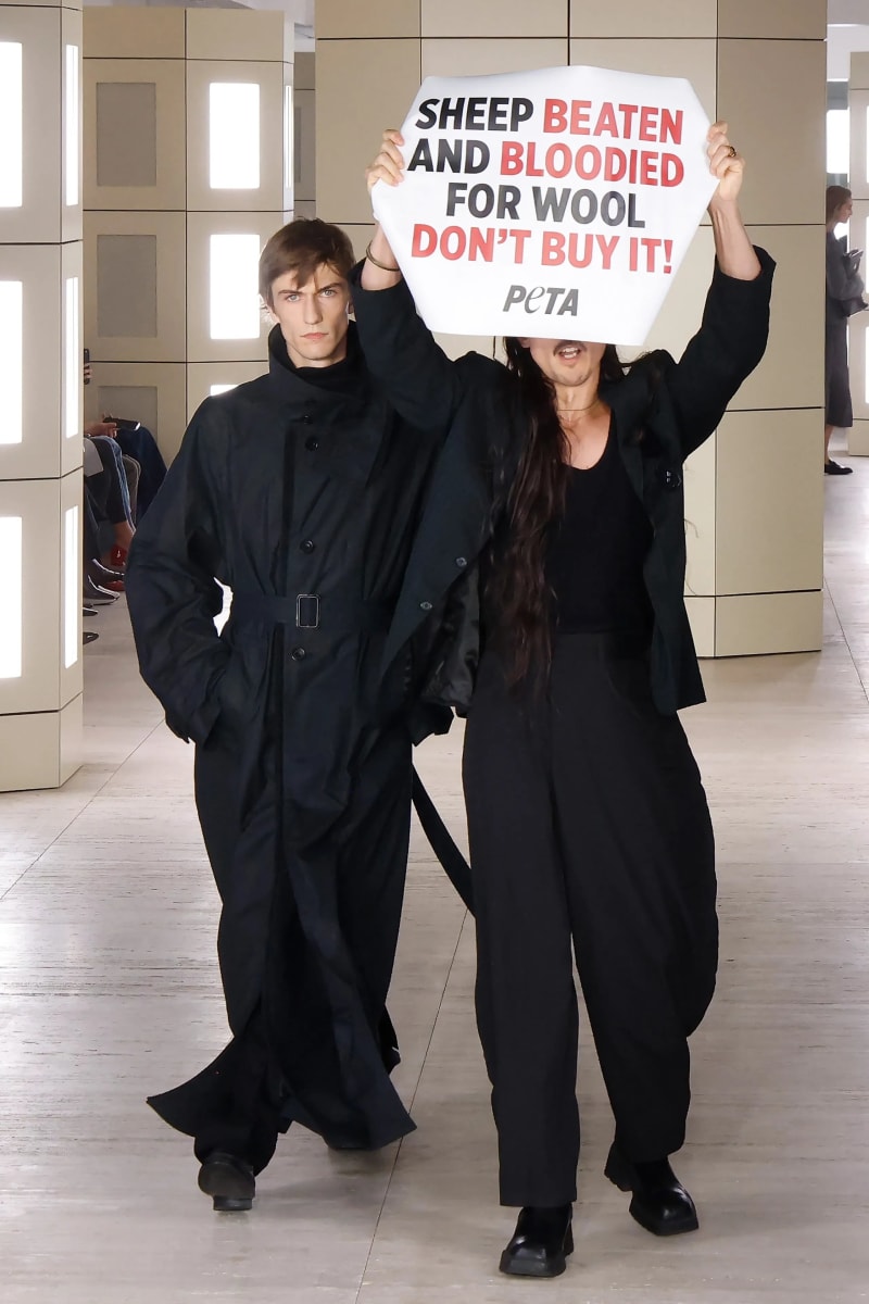 Man in black coat beside woman in black holding PETA protest sign, fashion runway.