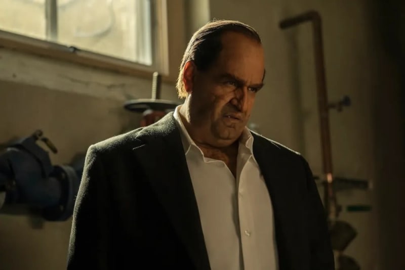 Man in black blazer and white shirt, scowling, in dim industrial room.
