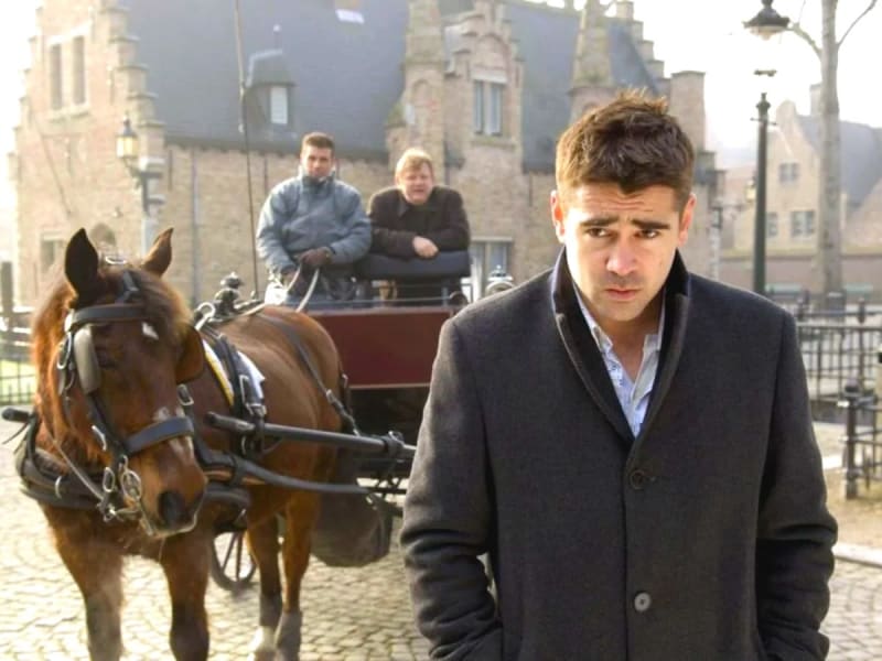Man in dark coat standing on cobblestone street, horse-drawn carriage and two men behind, European buildings in background.