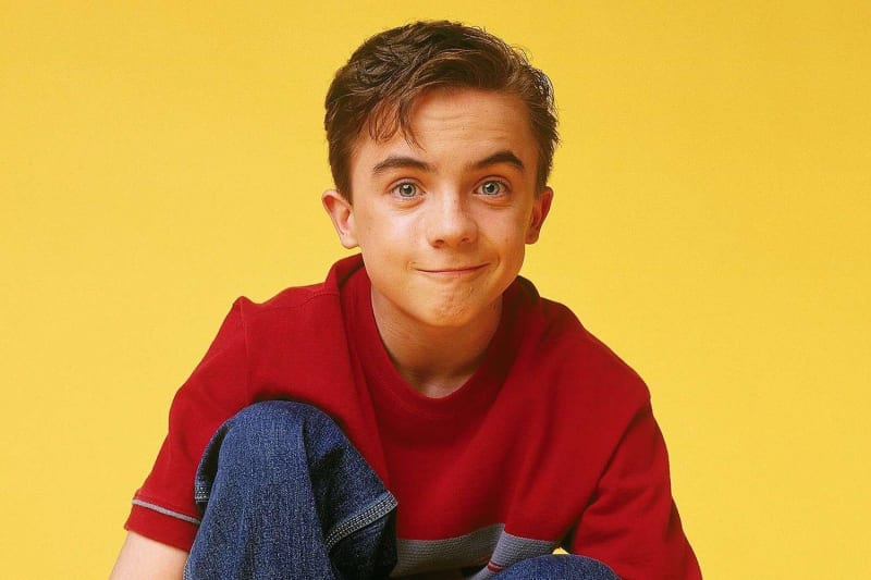 Boy in red t-shirt and jeans, seated smiling, yellow background.