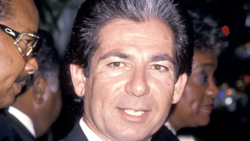 A man in a dark suit smiles at what appears to be a formal event, surrounded by other attendees in the background.