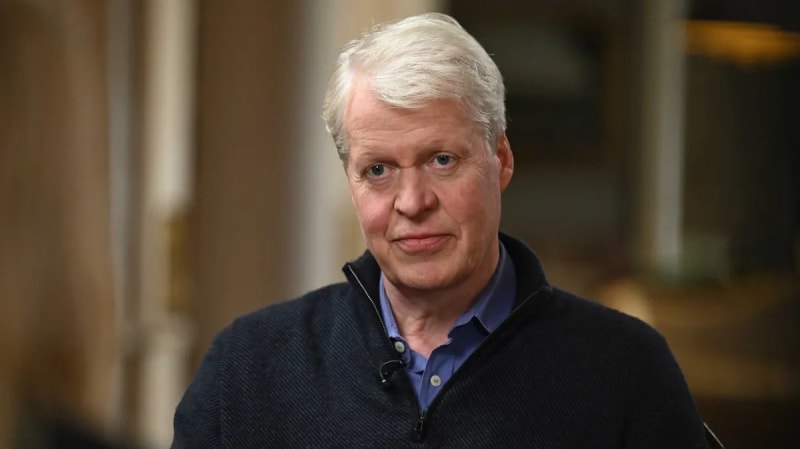 An older man with white hair wearing a dark cardigan over a blue shirt, looking at the camera in an indoor setting.