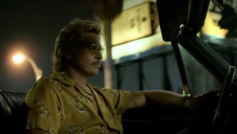 A man wearing sunglasses and a yellow patterned shirt sits in a car at night, hand on the steering wheel.