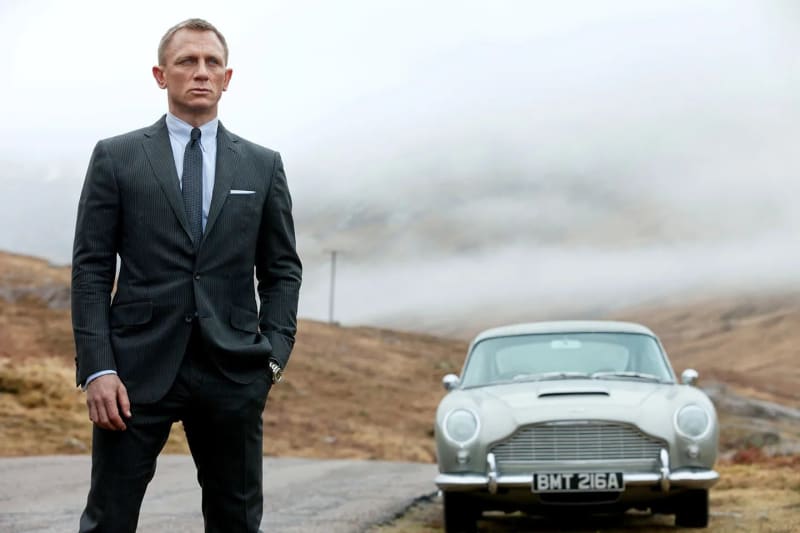 Man in dark pinstripe suit standing beside silver vintage car on misty highland road.