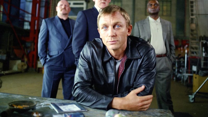 Man in black leather jacket leaning on table, three men in suits standing behind, warehouse setting.