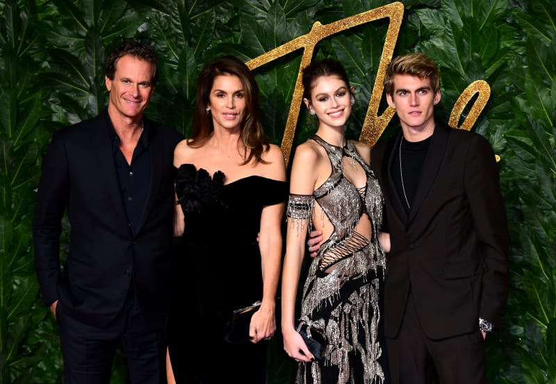 Four elegantly dressed people posing together at a formal event with a green foliage backdrop and gold arrow decoration.