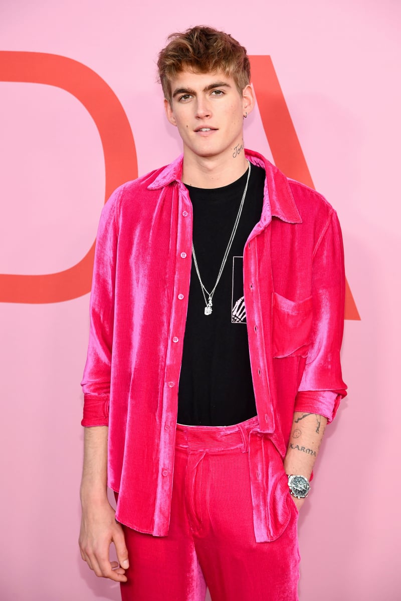 A young man wearing a bright pink velvet suit and black shirt poses against a pink background at an event.