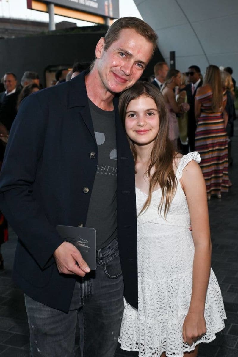 An adult and young girl pose together at an indoor event, with the man wearing a dark blazer and the girl in a white lace dress.