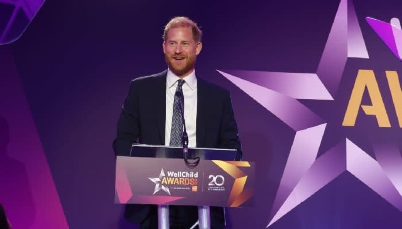 A man in a dark suit and tie speaking at a podium during the WellChild Awards ceremony with purple background.