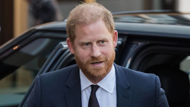 A man with reddish-blonde hair and beard wearing a dark suit and tie stands beside a dark vehicle.