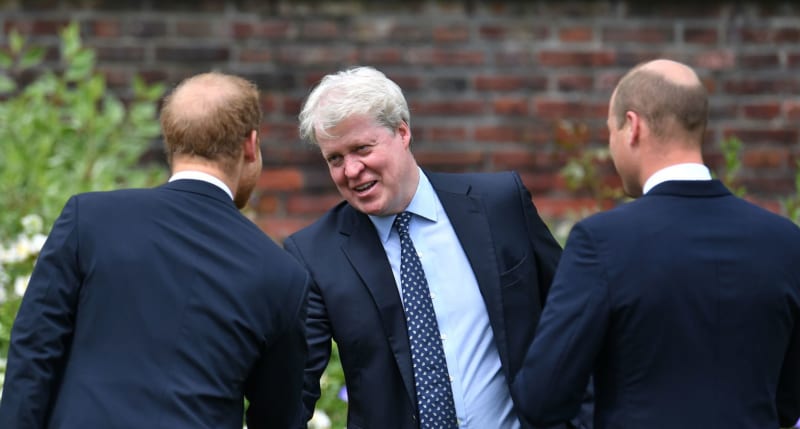 Three men in dark suits engaged in conversation against a brick wall backdrop.