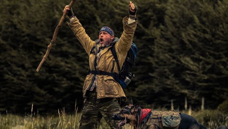 A bearded man in tan jacket and blue bandana celebrates triumphantly with arms raised while holding a walking stick, accompanied by a dog wearing a red pack in a forest setting.