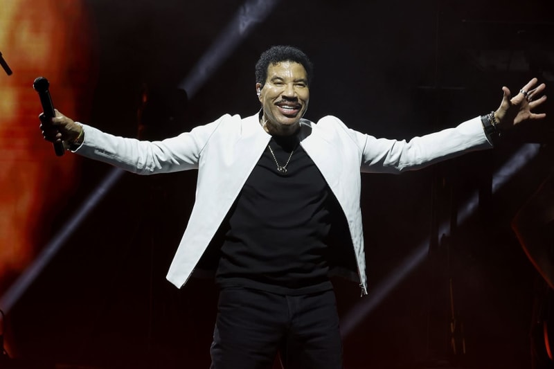 Man in white jacket and black outfit, arms outstretched holding microphone, on stage with red stage lighting.