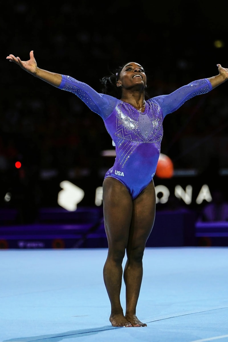 Woman in purple USA leotard, arms outstretched, on gymnastics floor.