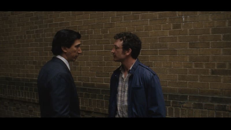 Two men in casual business attire stand facing each other against a brick wall in conversation.