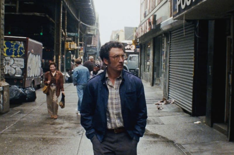 Man in blue jacket and plaid shirt, hands in pockets, walking on gritty urban street.