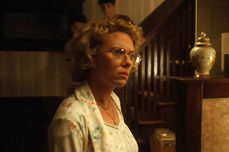 Woman in floral nightgown and glasses, looking worried, indoors near staircase.