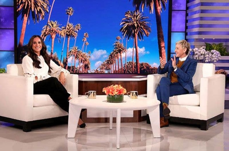 Two women sit in white chairs across from each other on a talk show set with a palm tree backdrop and flowers on the table between them.