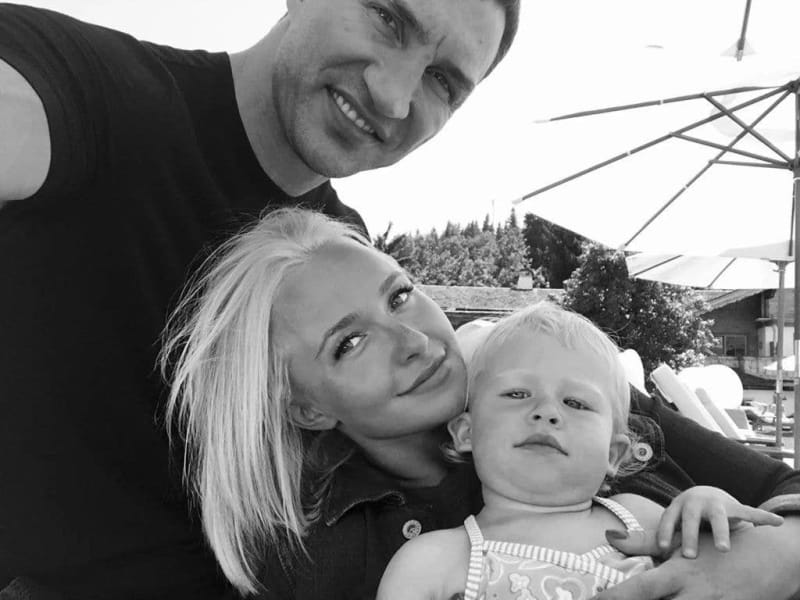 Black and white photo of a smiling man, woman with blonde hair, and young child posing together outdoors with mountains in background.
