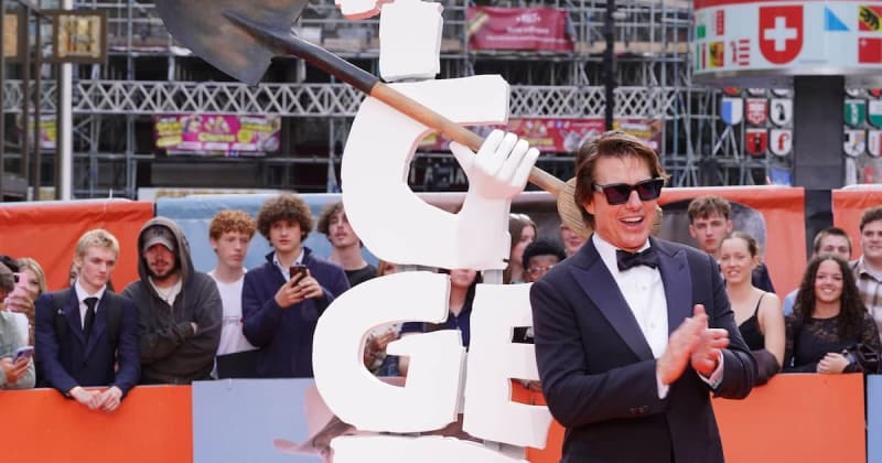 A man in a tuxedo and sunglasses holds a large white prop while posing at a red carpet event with crowds behind him.