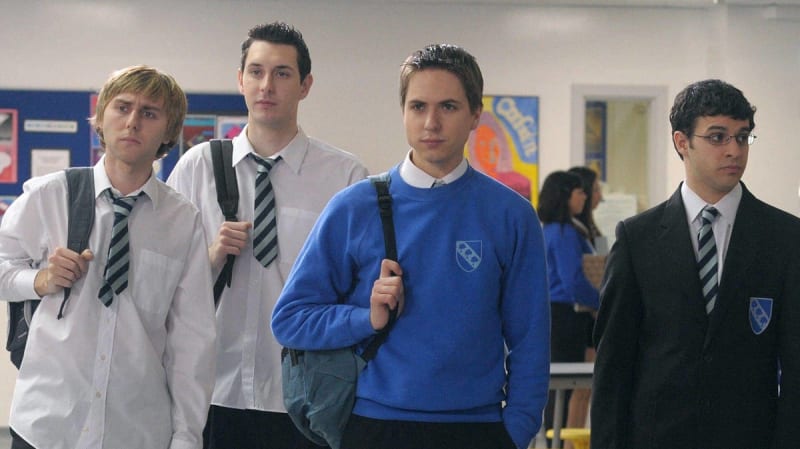 Four male students in school uniforms and backpacks stand in a hallway with colorful bulletin boards visible behind them.