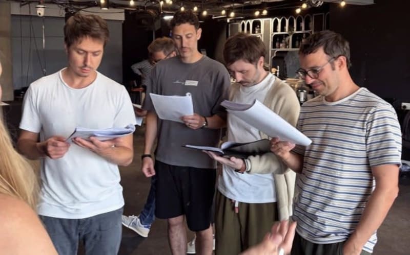 Four men examine papers and documents together in an indoor studio setting with warm lighting.