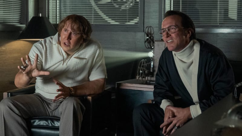 Two men sit in an office having a conversation, one gesturing while speaking and the other listening with a smile.