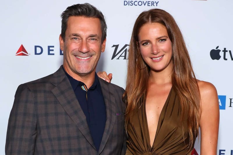 A man in a plaid blazer over a dark shirt stands beside a woman with long brown hair wearing a deep V-neck olive dress, posing together in front of a step-and-repeat banner featuring Delta, Variety, Apple TV, and Discovery logos.