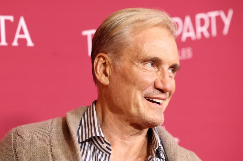 A man with light hair wearing a beige cardigan and striped shirt smiles at a red event backdrop.