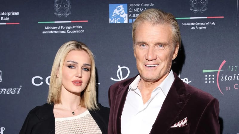 A young blonde woman in a cream top and black blazer stands beside an older silver-haired man in a dark burgundy velvet blazer and white dress shirt in front of a branded step-and-repeat backdrop featuring Italian government and cinema logos.