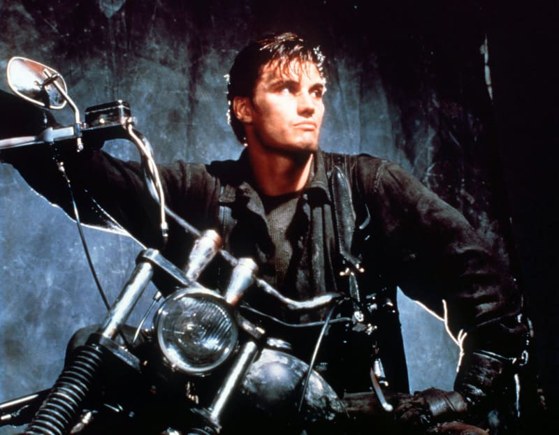 A man with slicked-back dark hair wearing a dark jacket sits astride a motorcycle, gripping the handlebars and gazing upward against a shadowy, textured backdrop.