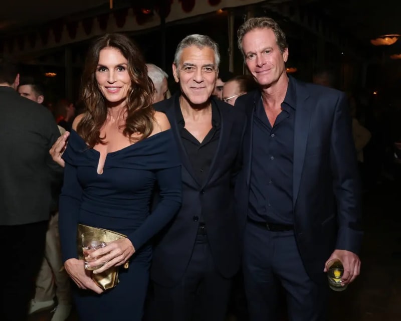 Woman in navy off-shoulder dress with gold clutch, man in black suit, man in navy suit, at indoor party.