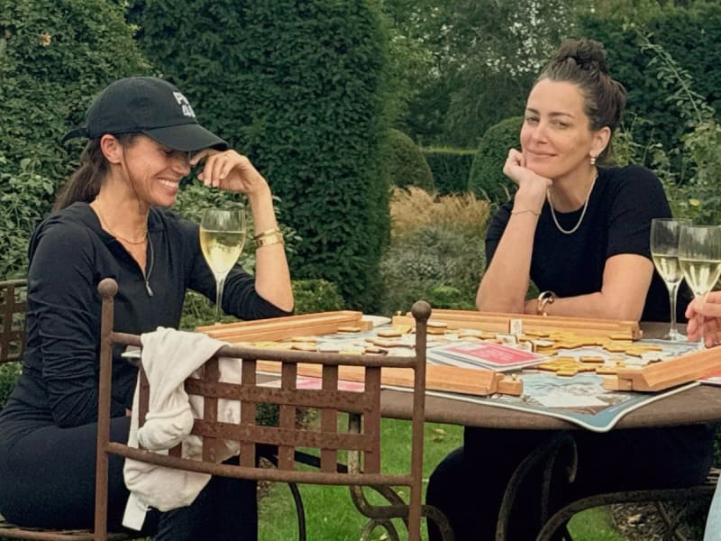 Two women sit at a wooden table in a garden, sharing wine and looking at documents, surrounded by green hedges.