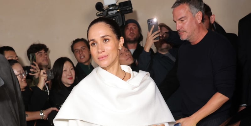 A woman in a white draped garment is photographed by media and onlookers at what appears to be a formal event or press gathering.