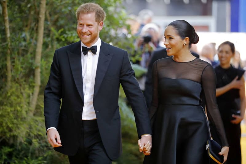 A man in a black tuxedo and a woman in a black dress walk hand-in-hand at a formal event with ivy-covered walls.