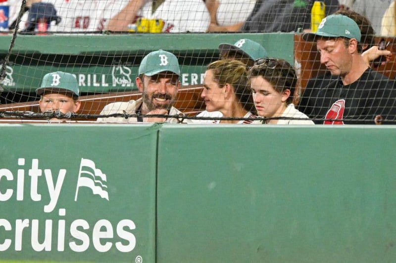 Group of people wearing Boston Red Sox caps watching from the dugout during a baseball game at Fenway Park.