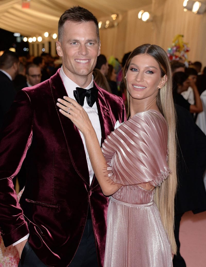 A couple at a formal event: man in burgundy velvet blazer with black bow tie, woman in blush pleated dress, both smiling.