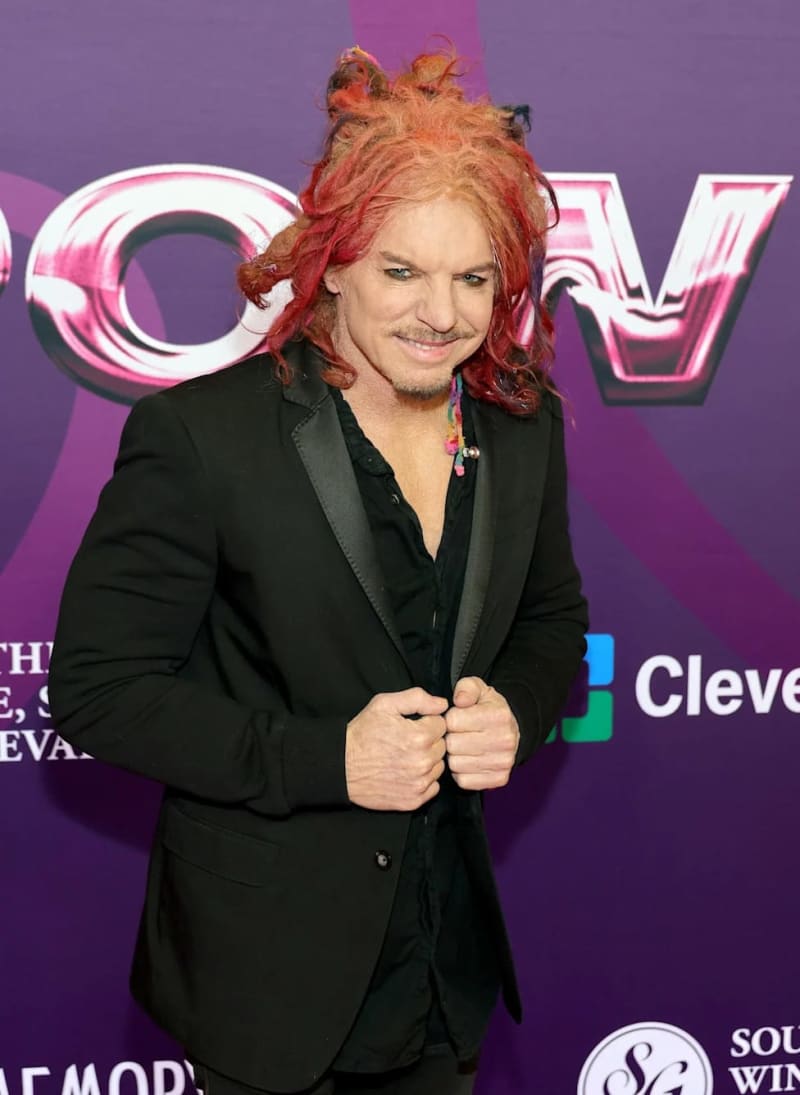 A person with voluminous red and blonde curled hair wearing a black blazer poses at a purple event backdrop.