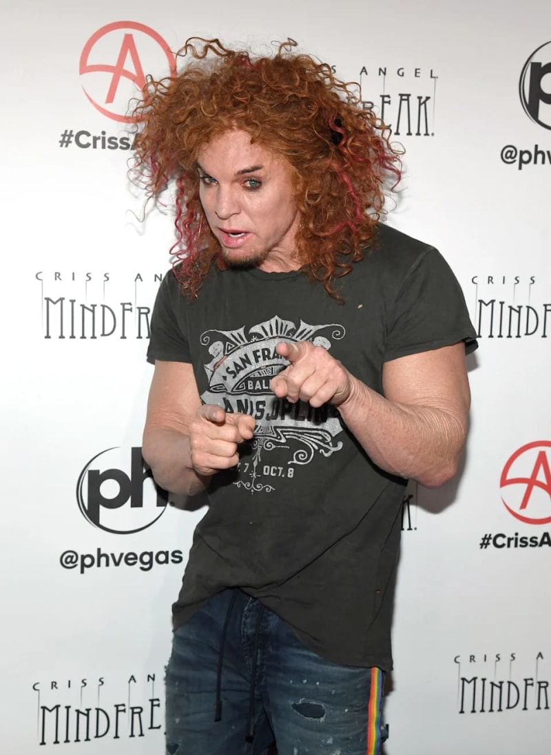 A person with curly reddish-brown hair wearing a black graphic t-shirt gestures while speaking at an event backdrop.