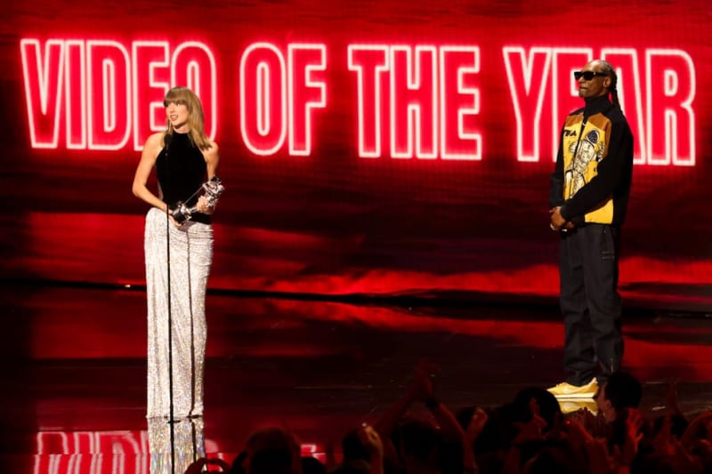Two presenters on stage at a Video of the Year awards ceremony with a glowing red neon backdrop and audience below.