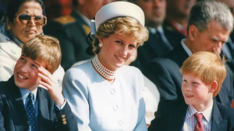 A woman in a white outfit and pearl necklace stands with two young boys in formal attire at what appears to be an official event.