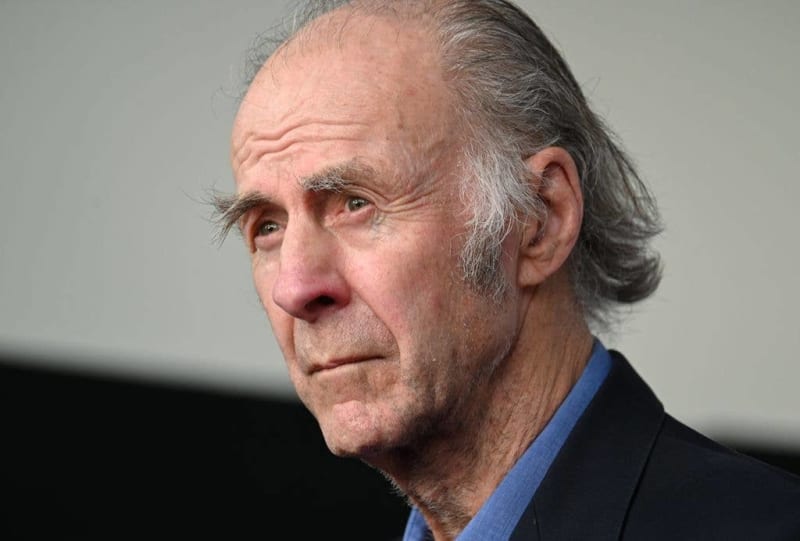 An older man with gray hair and facial hair wearing a dark jacket over a blue shirt, photographed in profile against a neutral background.