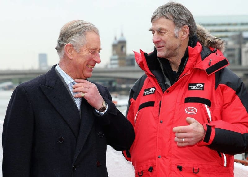 Two men in conversation on a rooftop with London skyline in background; one wearing formal dark coat, other in red and black jacket.