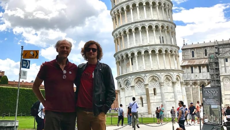 Two men in red shirts posing in front of the Leaning Tower of Pisa, Italy.